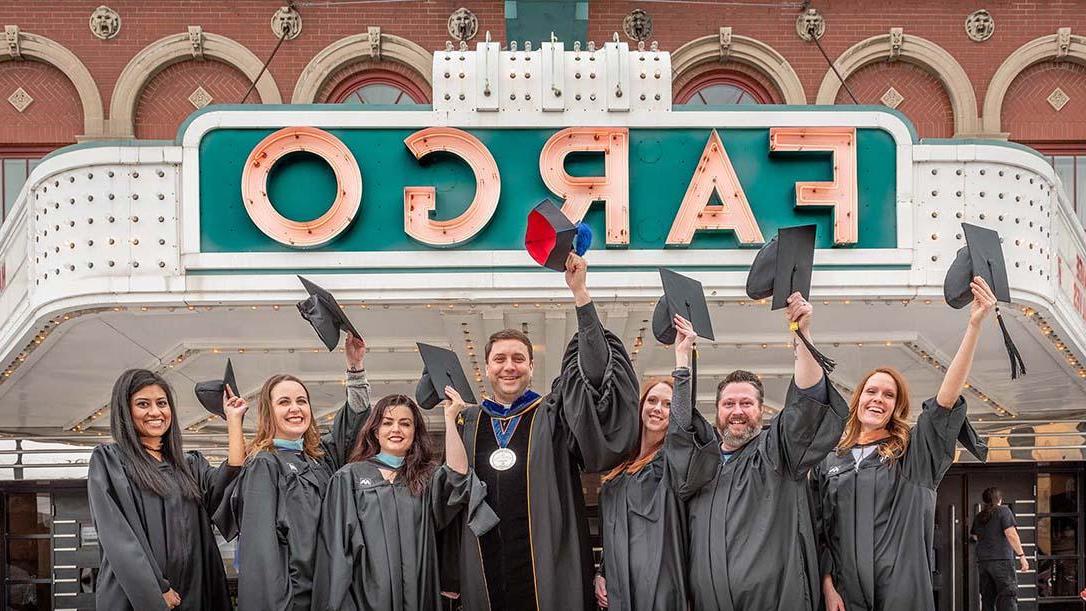Fargo adult students in their graduation gowns standing and waving their graduation caps outside the Fargo Theater sign with University of Mary President Monsignor Shea